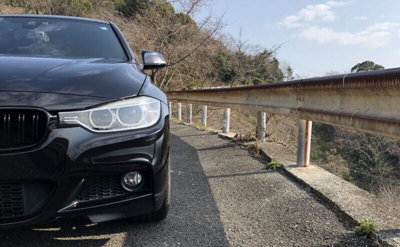 山道に停車した黒いBMW F30を左前方から撮影。ヘッドライトやキドニーグリル、フロントバンパーが見える