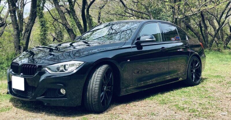 桜の木々を背景に黒いBMW F30を斜め前方から撮影。ブラックホイールやMスポーツのフロントデザインがよく分かる